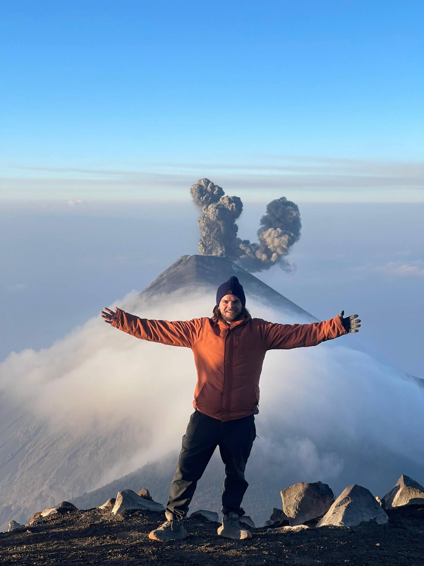 Erick con los brazos abiertos en la cima, frente a un volcán en erupción que sobresale de un mar de nubes